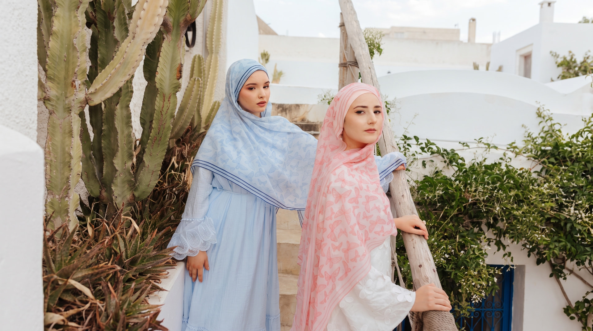 Two women in soft blue and pink butterfly-print Veiled Girl scarves on whitewashed steps