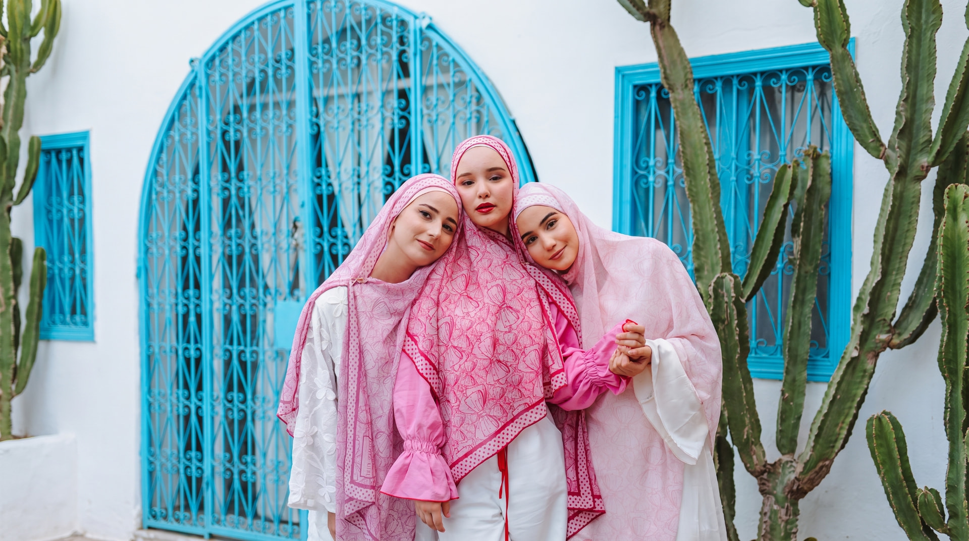 Three friends wearing pink printed Veiled Girl scarves in a blue-and-white Tunisian courtyard